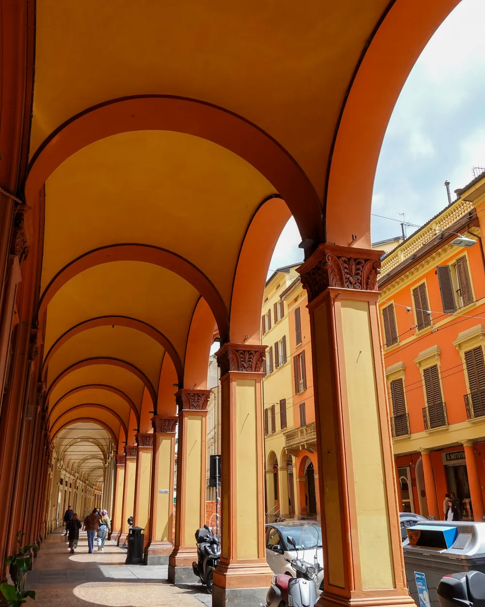 Bologna’da kemerli revakların altından geçen insanlar ve turuncu cepheli binalar
